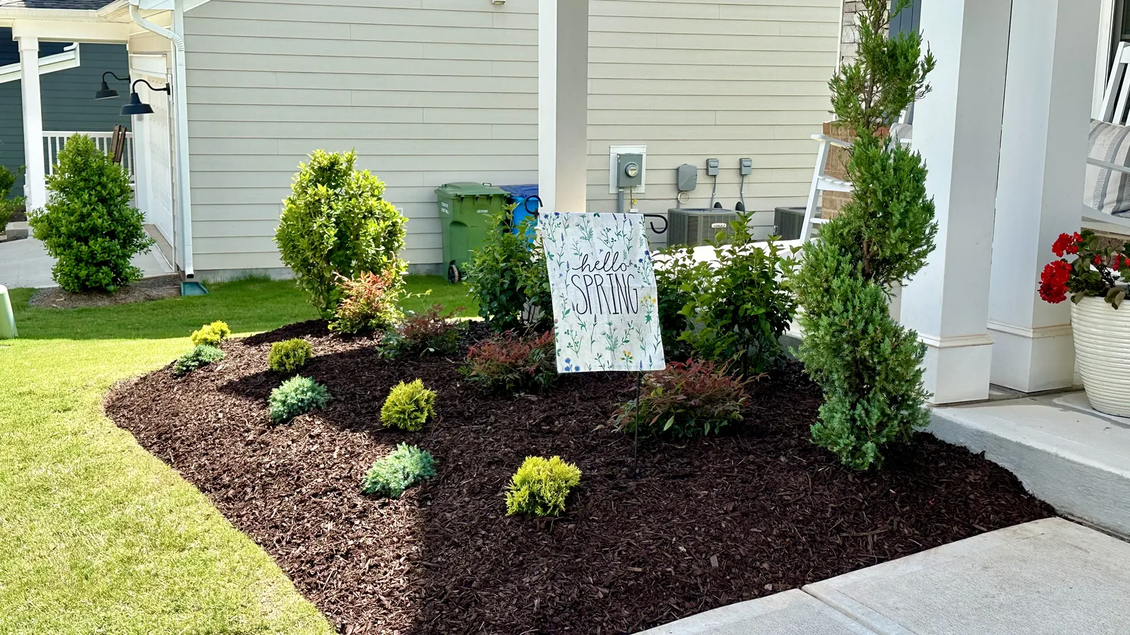 A mulched porch bed with mixed shrubs and a spring garden flag